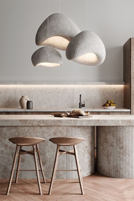 L'image montre une cuisine moderne avec un îlot central en béton brut, accompagné de deux tabourets en bois au design minimaliste. Au-dessus de l'îlot, trois suspensions aux formes organiques, rappelant des nuages, diffusent une lumière douce et accueillante. L'ensemble crée une atmosphère chaleureuse et contemporaine, alliant des matériaux naturels à un design épuré.