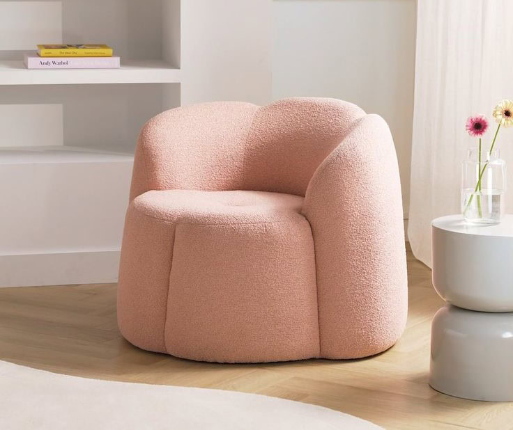 Fauteuil moderne et confortable en tissu rose pastel, avec un design arrondi. Placé sur un sol en bois clair, à côté d'une petite table blanche décorée avec des fleurs. En arrière-plan, une étagère blanche contenant des livres et des objets décoratifs. Une douce lumière naturelle éclaire la scène, créant une atmosphère chaleureuse et accueillante.