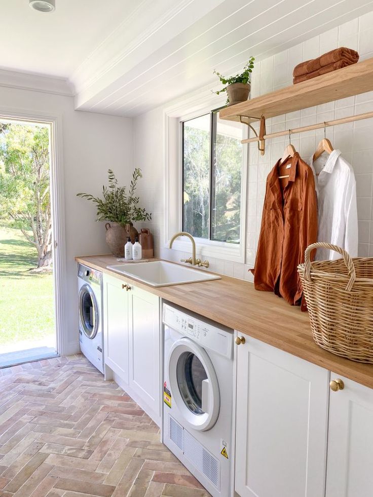 Buanderie lumineuse avec un plan de travail en bois clair, des machines à laver encastrées et une grande fenêtre donnant sur un jardin verdoyant. Une étagère ouverte au-dessus du plan de travail contient des paniers de rangement et des vêtements suspendus, créant un espace à la fois pratique et esthétique.