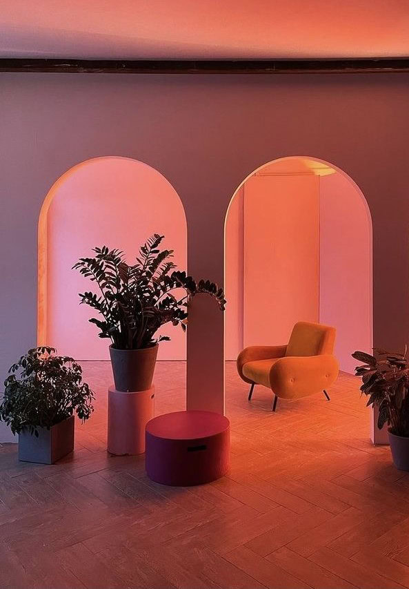 Un salon au design rétro avec trois arches illuminées par des lumières chaudes roses et oranges. Sous l'une des arches, un fauteuil orange crée un coin confortable. Des plantes d'intérieur sont disposées autour de la pièce, ajoutant une touche de verdure. Au premier plan, un pouf rose et un vase coloré complètent l'esthétique vibrante et accueillante de l'espace.