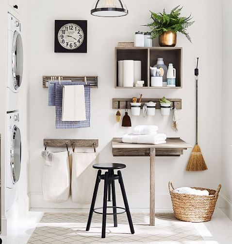 Un coin buanderie compact avec des machines à laver empilées à gauche, un petit bureau en bois avec un tabouret noir au centre, et des étagères murales pour le rangement de produits d'entretien. Des crochets suspendent des accessoires, et un panier en osier est posé au sol à droite, avec une balayette en bois accrochée au mur. Une horloge murale et une plante verte ajoutent une touche décorative à l'ensemble.