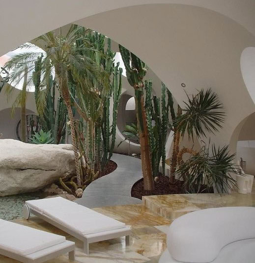 Un espace intérieur moderne intégrant un jardin tropical avec des plantes exotiques telles que des palmiers et des cactus, des rochers décoratifs, des arches blanches et des formes courbes. Au premier plan, deux chaises longues blanches sont disposées sur un sol en pierre naturelle, créant une atmosphère relaxante et harmonieuse.